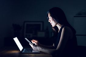 Busy young woman text messaging on smartphone while working on laptop till late