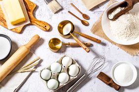 Top view of various bakery ingredients and items such as eggs, butter, flour, sugar, rolling pin, measure cups, hand whisk and serving scoop disposed all around the image on a bakery concept background